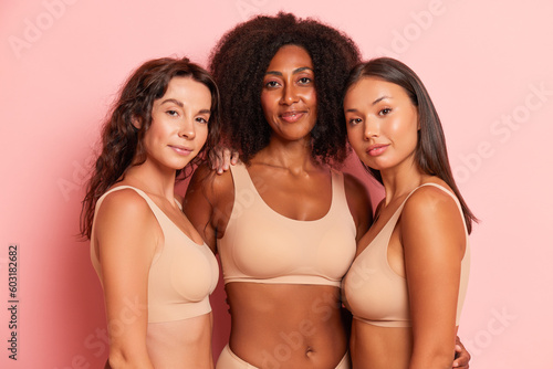 Three cheerful girlfriends pose against pink wall wearing beige sport tops, one black-skinned girl with her friends by her sides, happy time concept, copy space © South House Studio