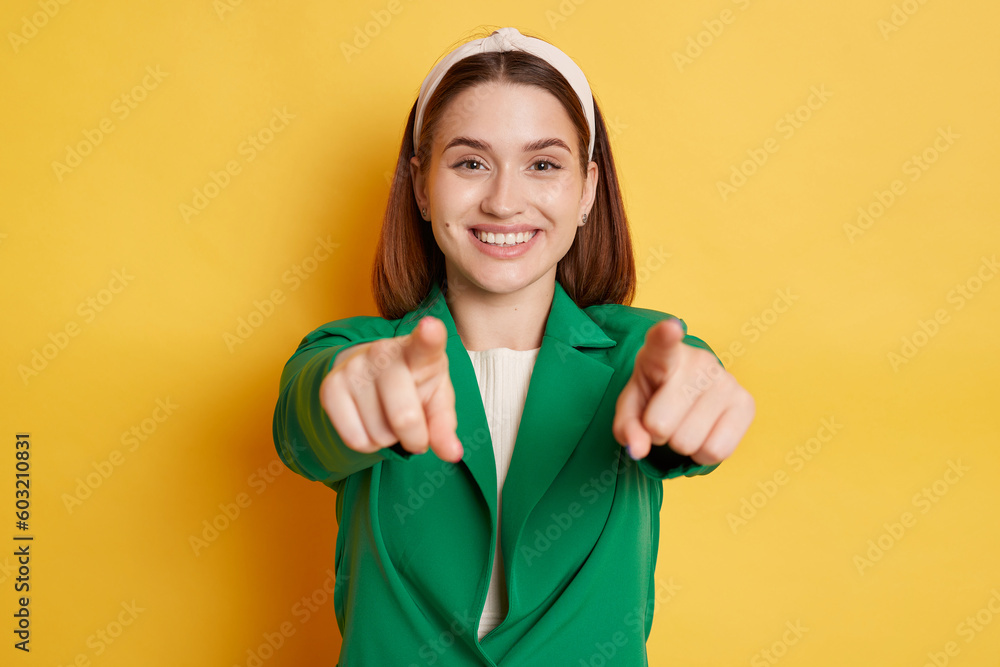 Joyful smiling woman wearing green jacket posing isolated over yellow background pointing to camera choosing you positive reaction. Joyful smiling woman wearing green jacket posing isolated over yellow background pointing to camera choosing you positive reaction.