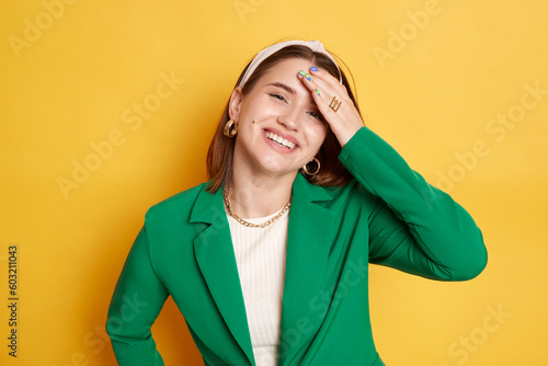 Beautiful delighted woman wearing green jacket keeps hand on forehead smiling remembering forget something posing isolated over yellow background.