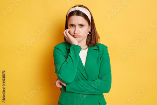 фотографія Bored sad offended woman wearing green jacket posing isolated over yellow background keeps hand under chin looking at camera with unhappy face displeased emotions