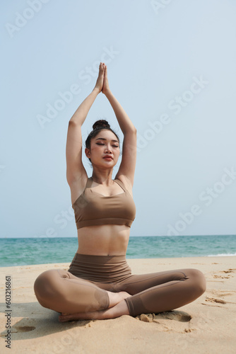 Young woman sitting in lotus position when her arms above head when meditating on beach