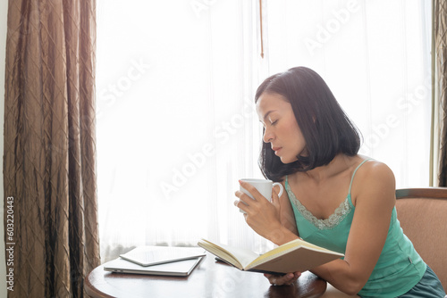 Young woman at home sitting on modern chair in front of window relaxing in her living room reading book and drinking coffee or tea..Beautiful young woman sitting by the window. woman enjoying book.