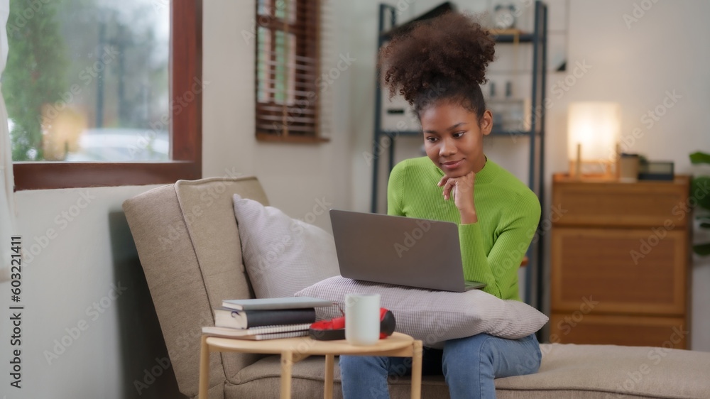 Smiling woman using laptop sitting on the sofa at home. Beautiful young black female shopping or chatting online in social networks, having fun, watching movie, freelance working on laptop projects. Smiling woman using laptop sitting on the sofa at home. Beautiful young black female shopping or chatting online in social networks, having fun, watching movie, freelance working on laptop projects.