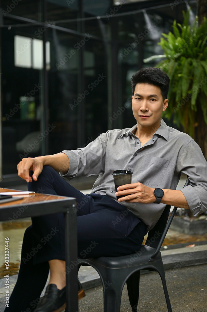 Smiling millennial Asian man sits at a coffee shop outdoor space with his afternoon coffee. Smiling millennial Asian man sits at a coffee shop outdoor space with his afternoon coffee.