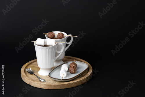 Two beautiful white porcelain cups of hot Chocolate drink with marshmallows and chocolates skewers over wooden table on black background. Winter time. Holiday concept..