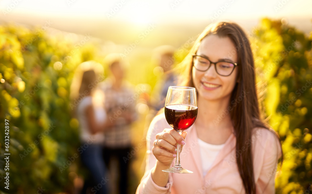 Portrait of a young, millennial woman holding a glass of organic bio red wine outdoors in a vineyard with her friends in the background - Vine-growing, and wine-tasting concept Portrait of a young, millennial woman holding a glass of organic bio red wine outdoors in a vineyard with her friends in the background - Vine-growing, and wine-tasting concept