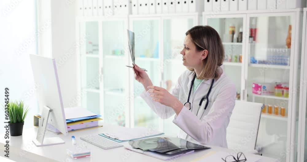 Focused doctor in work uniform examines X-ray picture of bone fracture sitting at table in clinic office. Female employee looks at radiograph slow motion Focused doctor in work uniform examines X-ray picture of bone fracture sitting at table in clinic office. Female employee looks at radiograph slow motion