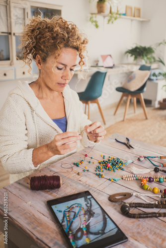 Woman at home making beads jewels using instructions online on a tablet. Alternative job homemade workplace. Adult female create bracelets and necklaces as artisan. New business and hobby leisure photo