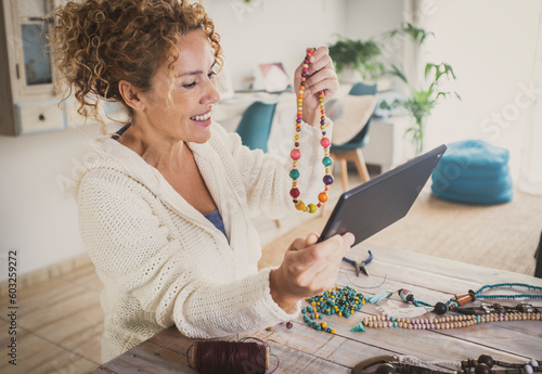 Happy woman indoor leisure hobby work activity producing beads accessories necklaces showing her jewel in video call using tablet and wireless connection technology. New business at home lifestyle photo
