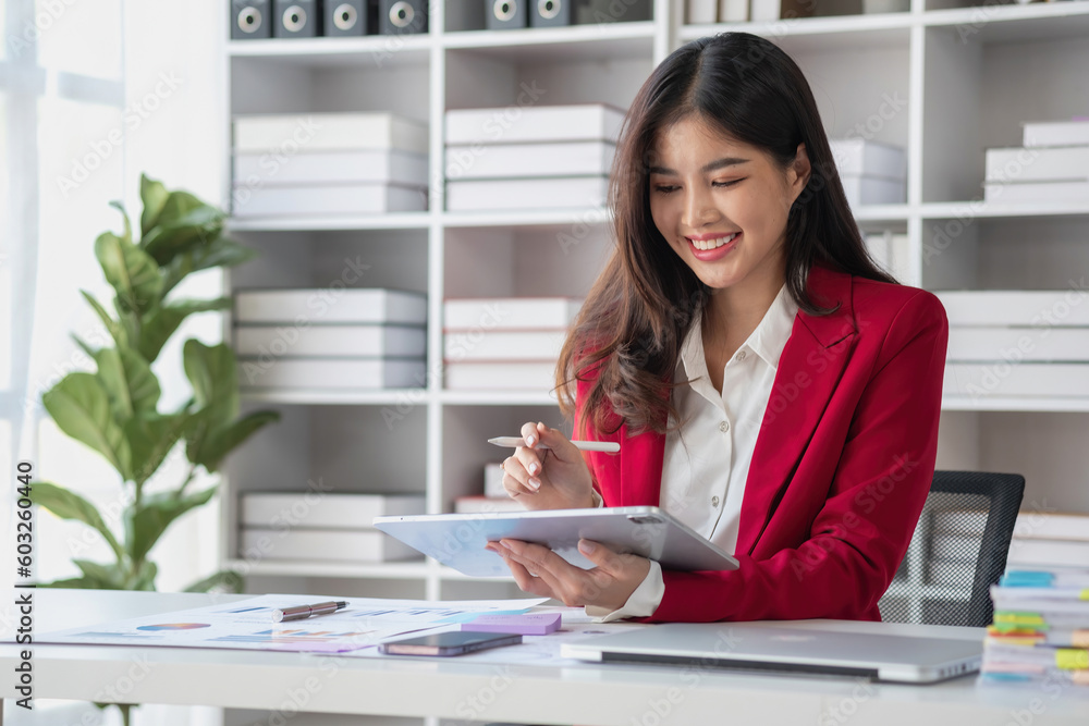 Asian businesswoman in red suit calculating company sales with calculator, laptop, and tablet on table Interior of businesswoman office at the modern workstation. Asian businesswoman in red suit calculating company sales with calculator, laptop, and tablet on table Interior of businesswoman office at the modern workstation.