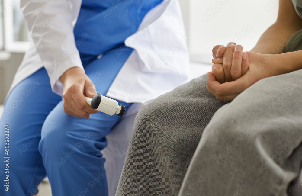 Neurologist with medical hammer examines child patient. Female doctor in scrubs checks knee jerk reflex of teenage school girl in gray pants. Cropped shot. Neurology, health checkup concept background