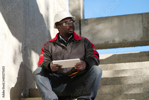 Tired african american worker sits at construction site and holds tablet PC