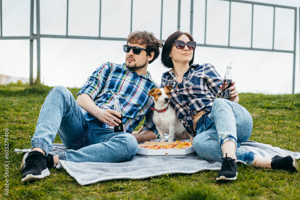 Young family with their dog sitting on the grass and eating pizza snack outdoor