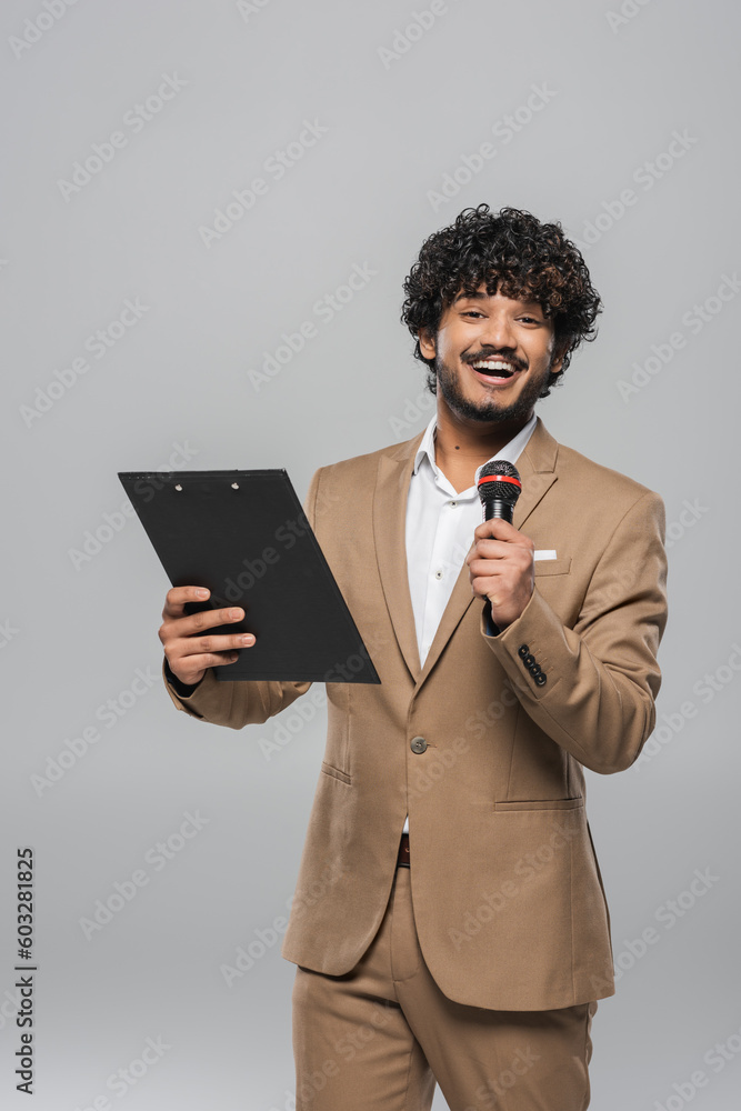 Cheerful and curly young indian host of event holding microphone and clipboard while looking at camera during celebration isolated on grey Cheerful and curly young indian host of event holding microphone and clipboard while looking at camera during celebration isolated on grey