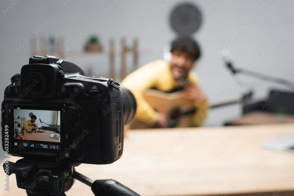 screen of digital camera on tripod standing near blurred young indian podcaster holding acoustic guitar near microphone and laptop on table in podcast studio