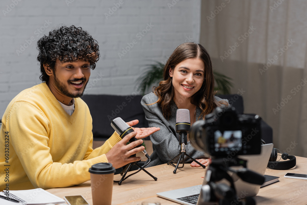 curly indian man in yellow jumper talking in microphone and pointing at charming colleague near laptop, coffee to go and smartphones in front of blurred digital camera in broadcasting studio curly indian man in yellow jumper talking in microphone and pointing at charming colleague near laptop, coffee to go and smartphones in front of blurred digital camera in broadcasting studio