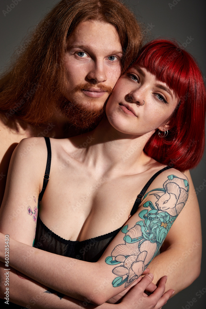 Studio portrait of a sexy half-naked couple in love. A handsome man with long hair and a beard gently hugs his woman with red hair, a piercing and a tattoo. Love, passion and tenderness. Studio portrait of a sexy half-naked couple in love. A handsome man with long hair and a beard gently hugs his woman with red hair, a piercing and a tattoo. Love, passion and tenderness.