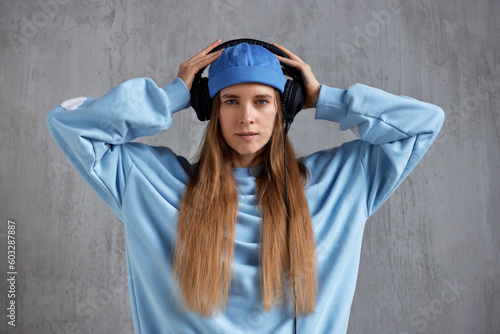 A young pretty long-haired girl in a blue sweater and cap wears DJ headphones on her head and holds them with her hands. Music and beauty. photo