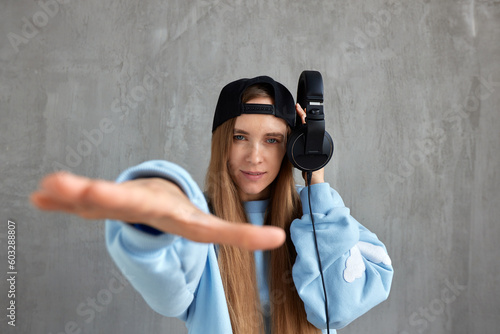 A young pretty long-haired DJ girl in a blue sweater and a black baseball cap holds headphones in her hand and imitates the movements of a hip-hop musician. Music, fun and beauty.