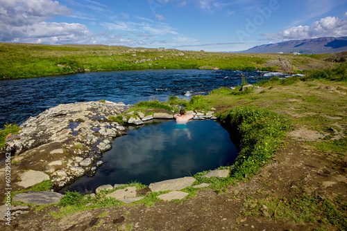 Man relaxing and bathing in natural geothermal heated hot pool, hot spring in Iceland in summer. Green rolling hills and blue sky on background. Hot spring is called Fosslaug hot pot. photo