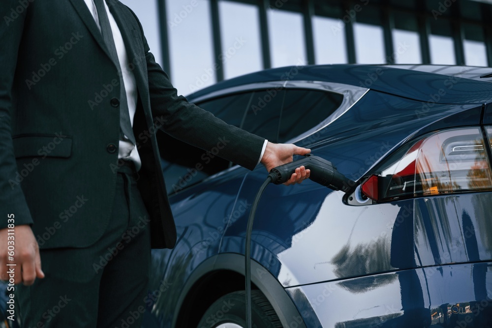 Close up view, putting charger into the socket. Businessman is standing near his electric car outdoors Close up view, putting charger into the socket. Businessman is standing near his electric car outdoors