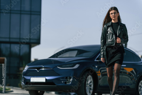 In black jacket and skirt. Young woman is near her electric car outdoors © standret