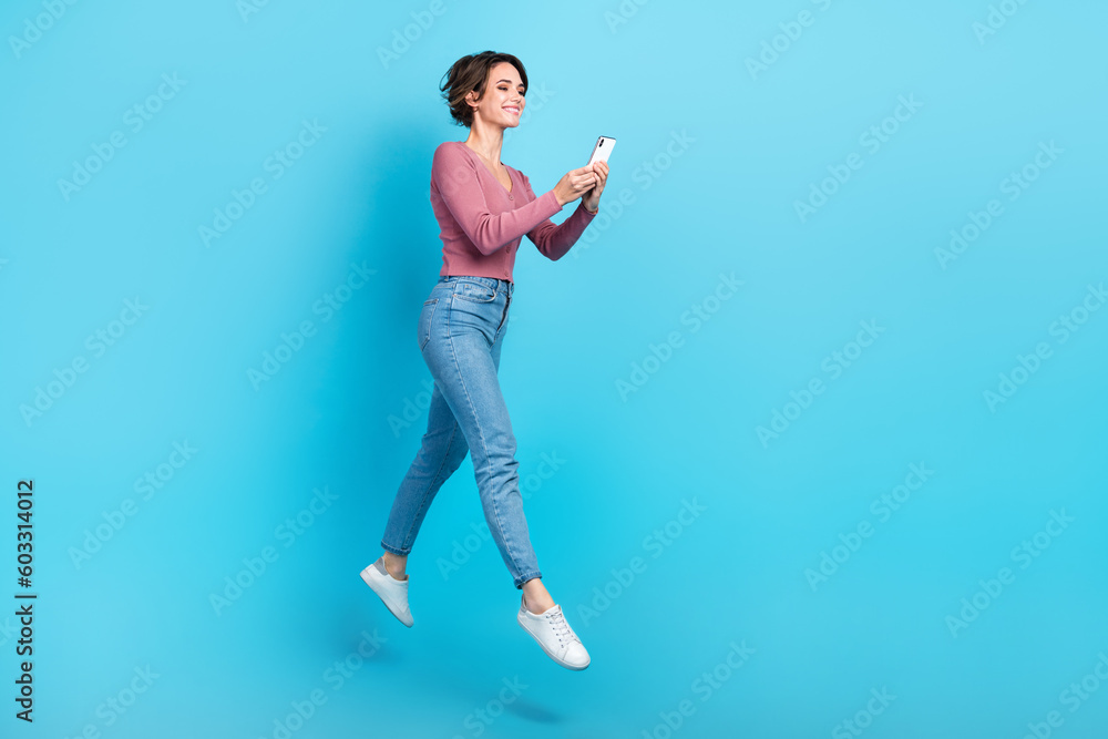 Full size profile portrait of active charming girl jumping use telephone chatting isolated on blue color background Full size profile portrait of active charming girl jumping use telephone chatting isolated on blue color background