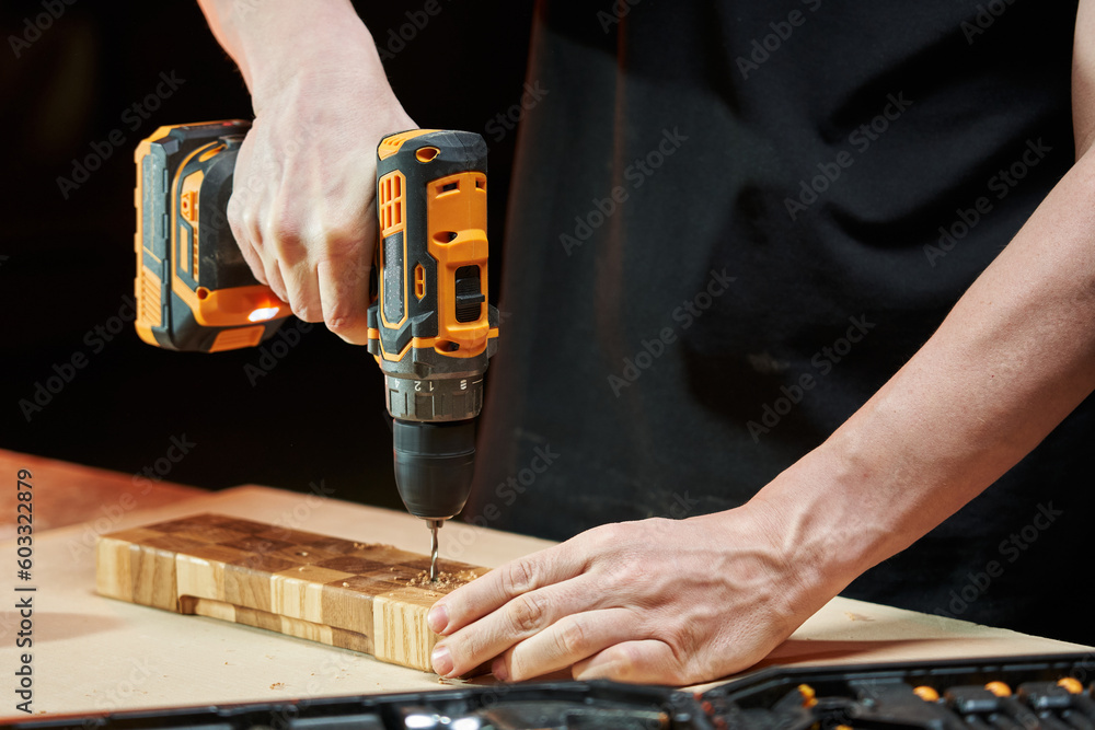 Hands of caucasian worker drilling a wooden detail with a cordless electric drill-driver on a wooden workbench