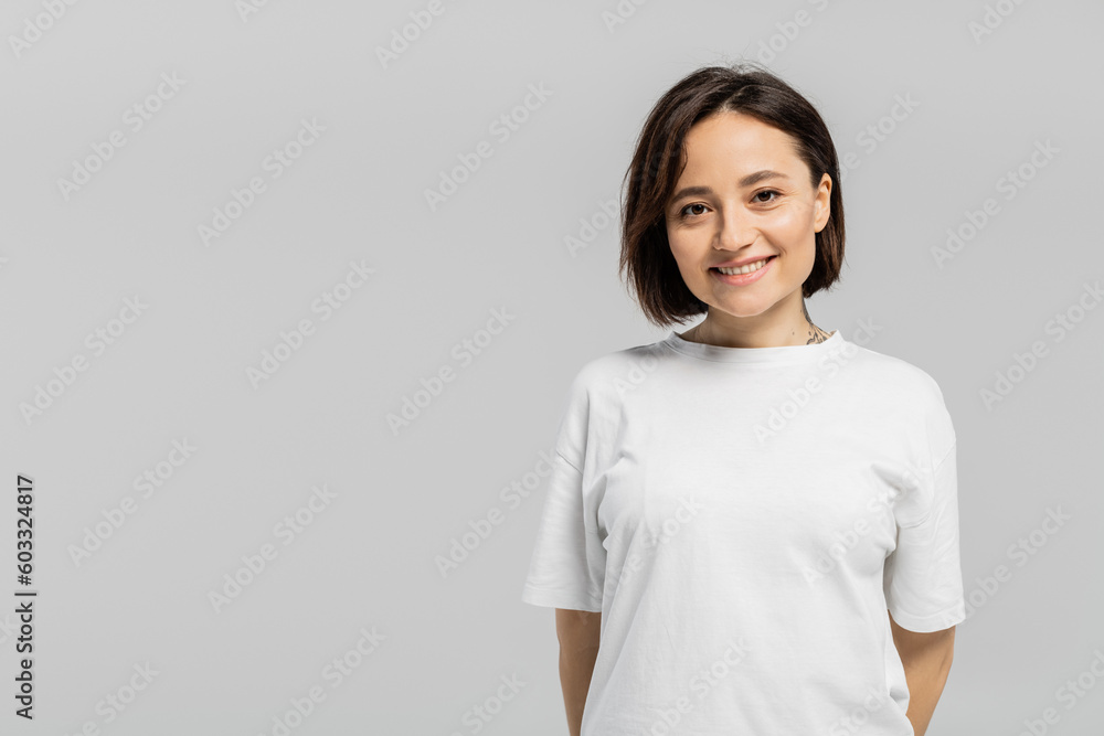 happy and tattooed woman with short hair and natural makeup standing in white t-shirt and smiling while looking at camera isolated on grey background with copy space happy and tattooed woman with short hair and natural makeup standing in white t-shirt and smiling while looking at camera isolated on grey background with copy space