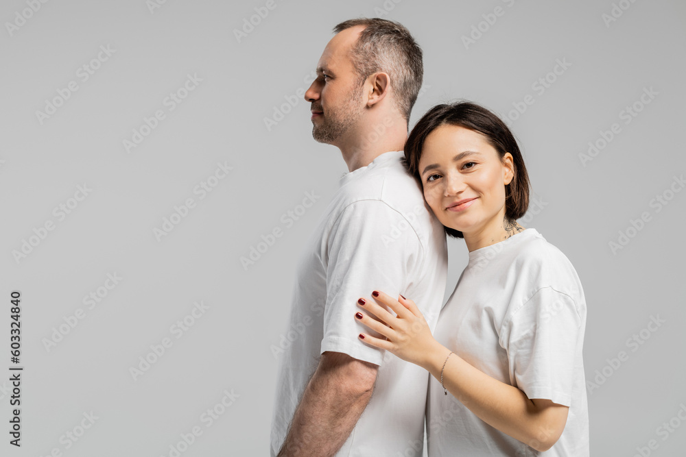 tattooed woman with short brunette hair leaning on back of husband while standing together in white t-shirts and looking at camera isolated on grey background, happy couple tattooed woman with short brunette hair leaning on back of husband while standing together in white t-shirts and looking at camera isolated on grey background, happy couple