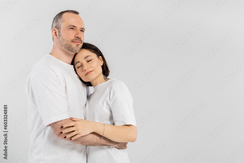 tattooed woman with short brunette hair and closed eyes leaning on chest of husband with bristle while standing together in white t-shirts isolated on grey background, happy couple tattooed woman with short brunette hair and closed eyes leaning on chest of husband with bristle while standing together in white t-shirts isolated on grey background, happy couple