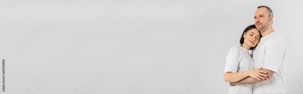 tattooed woman with short brunette hair leaning on chest of husband while standing together in white t-shirts isolated on grey background, happy couple, banner tattooed woman with short brunette hair leaning on chest of husband while standing together in white t-shirts isolated on grey background, happy couple, banner