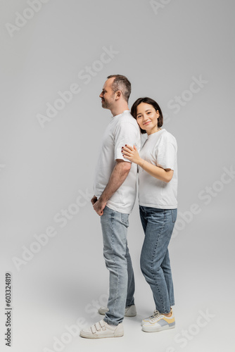 full length of tattooed woman with short brunette hair leaning on back of smiling husband while standing together in white t-shirts and denim jeans on grey background, happy couple