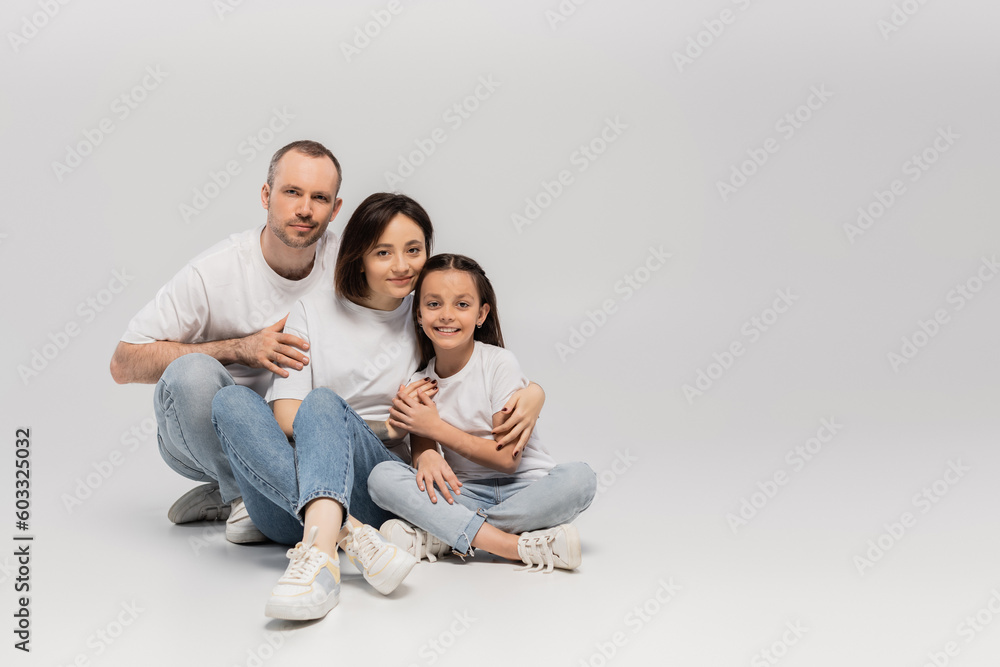joyous father and mother with short hair hugging cheerful preteen daughter while sitting with crossed legs in white t-shirts and blue denim jeans on grey background, Happy children's day joyous father and mother with short hair hugging cheerful preteen daughter while sitting with crossed legs in white t-shirts and blue denim jeans on grey background, Happy children's day