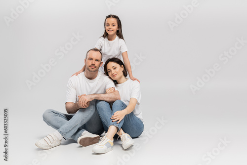 joyous preteen girl hugging mother and father in white t-shirts and blue denim jeans while bonding together and looking at camera on grey background, Happy children's day