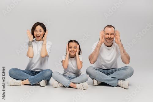 amazed father and tattooed mother with short hair and excited preteen daughter sitting with crossed legs in white t-shirts and blue denim jeans on grey background, Happy children's day