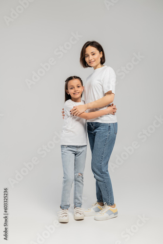 full length of mother with short hair and tattoo on hand hugging preteen daughter while standing together in white t-shirts and blue denim jeans on grey background, International child protection day