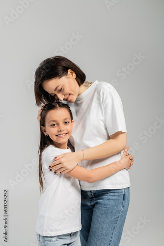 happy mother with short hair and tattoo on hand hugging preteen daughter while standing together in white t-shirts and blue denim jeans on grey background, International child protection day © LIGHTFIELD STUDIOS