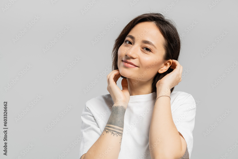 alluring and tattooed woman touching her short hair and standing in white t-shirt, smiling while looking at camera isolated on grey background with copy space alluring and tattooed woman touching her short hair and standing in white t-shirt, smiling while looking at camera isolated on grey background with copy space