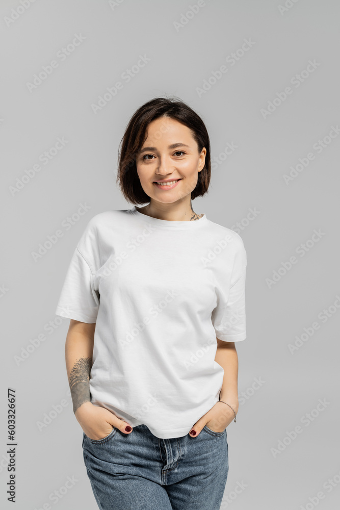 alluring and tattooed woman with short hair and natural makeup standing in white t-shirt and posing with hands in pockets of blue denim jeans while looking at camera isolated on grey background alluring and tattooed woman with short hair and natural makeup standing in white t-shirt and posing with hands in pockets of blue denim jeans while looking at camera isolated on grey background