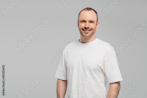 unshaved happy man with bristle standing in white t-shirt and looking at camera while posing isolated on grey background in studio, copy space, happiness