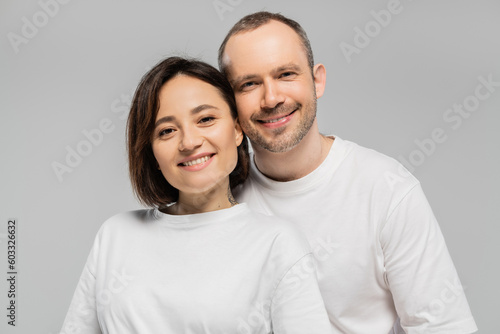 cheerful husband and tattooed wife with short brunette hair standing together in white t-shirts and looking at camera isolated on grey background in studio, happy couple