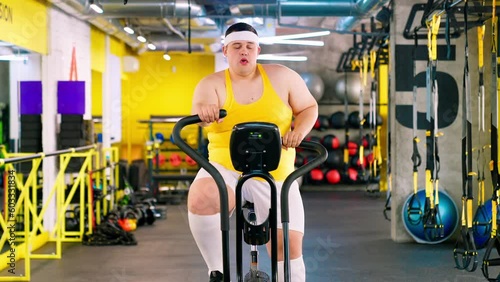 The fat man is exercising in the gym and looking at the burger and pizza. Obesity of a young man and doing sports in the gym. The fat man eats a burger. photo