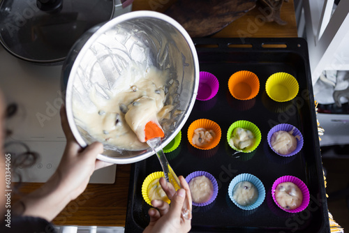 making of delicious cupcake in kitchen. Muffin tin with liner, ingredients, and utensils are seen. A woman is adding mixture into the holes