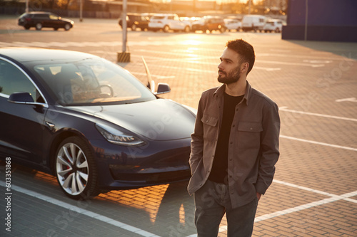 Evening time. Beautiful sunlight. Man is standing near his electric car outdoors