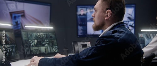 Male security officer uses tablet and controls CCTV cameras with AI facial recognition in police surveillance center. PC monitors and big digital screens showing security cameras footage. Monitoring.