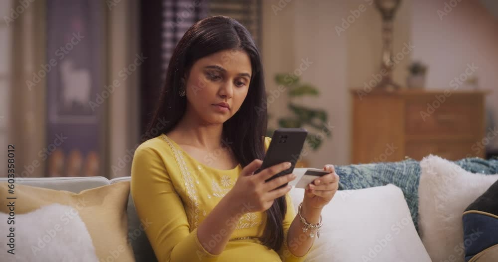 Portrait of Indian Woman Utilizing Smartphone and Credit Card for Online Purshases at Home: Paying Bills, Purchasing Stylish Clothing, Securing a Vacation Deal; Online Shopping, Ecommerce. Medium Arc Portrait of Indian Woman Utilizing Smartphone and Credit Card for Online Purshases at Home: Paying Bills, Purchasing Stylish Clothing, Securing a Vacation Deal; Online Shopping, Ecommerce. Medium Arc