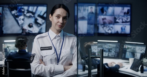 Female security worker in police surveillance center looks at camera. Male employees work on background. CCTV cameras footage displayed on big digital screen. Tracking and monitoring system. Portrait.