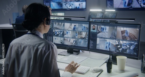 Security officer in headset controls CCTV cameras with AI facial recognition on PC. Female employee works in security control center. Computer monitors showing surveillance cameras video footage.
