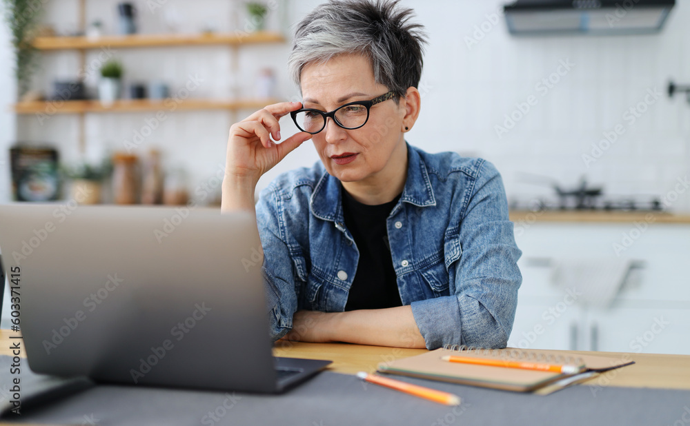 Confused mature woman in front of a laptop at the workplace, made a mistake. - obrazy, fototapety, plakaty 